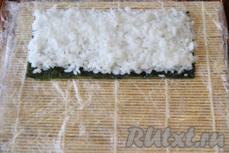 Лист нори разрезать на две части. На циновку выложить лист нори блестящей стороной вниз, затем выложить часть риса и разровнять его по поверхности листа нори. Оставить пустым дальний от вас край шириной 1 см. Чтобы рис не прилипал к рукам нужно руки смочить в подкисленной воде (в воду добавить пару ложек лимонного сока). 