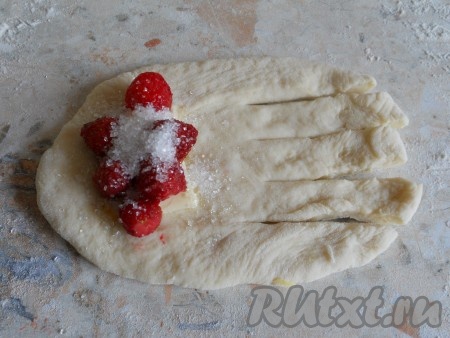 Далее разделить дрожжевое тесто на 15 частей. Каждую часть теста раскатать в небольшую овальную лепёшку. На край лепёшки поместить по несколько ягод клубники (лучше использовать мелкие ягодки), посыпать 1 чайной ложкой сахара. Другой край лепёшки разрезать вдоль на 4-5 полос.