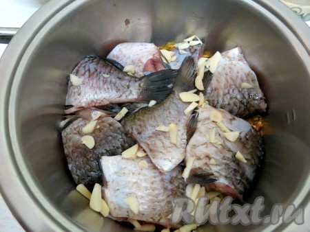 На дно кастрюли с толстым дном наливаем 2 столовые ложки растительного масла, выкладываем половину обжаренных овощей, добавляем рыбу, посыпаем чесноком, нарезанным на пластины.
