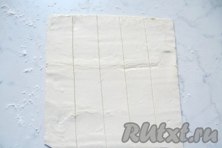 Размороженное слоёное тесто на столе, присыпанном мукой, немного раскатать, затем нарезать его на полоски шириной 3-4 сантиметра.